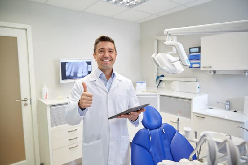 eople, medicine, stomatology and healthcare concept - happy middle aged male dentist in white coat with tablet pc computer showing thumbs up at dental clinic office